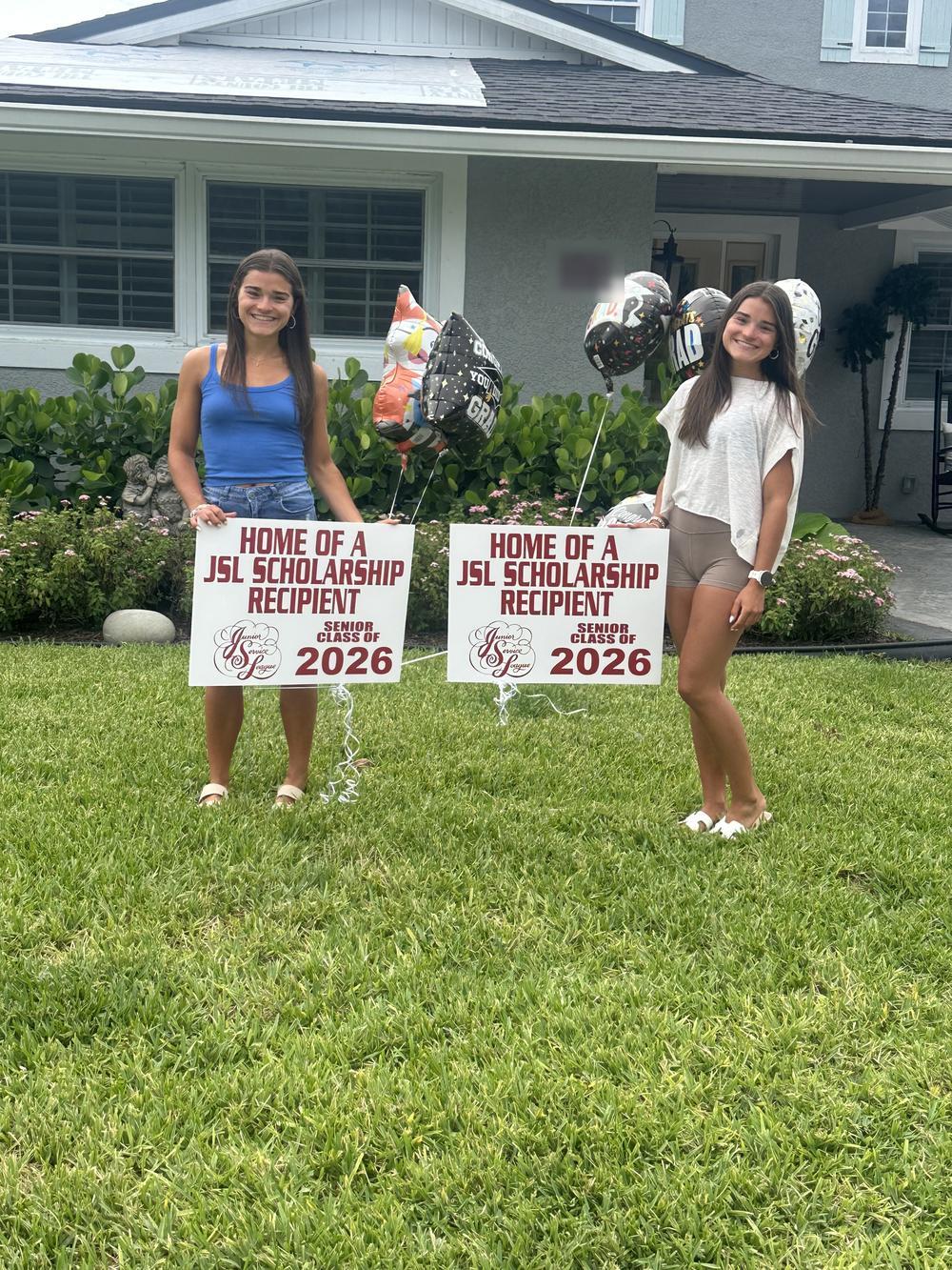 Two graduating seniors stand in a front yard with their Home of a JSL Scholarship Recipient signs and graduation balloons