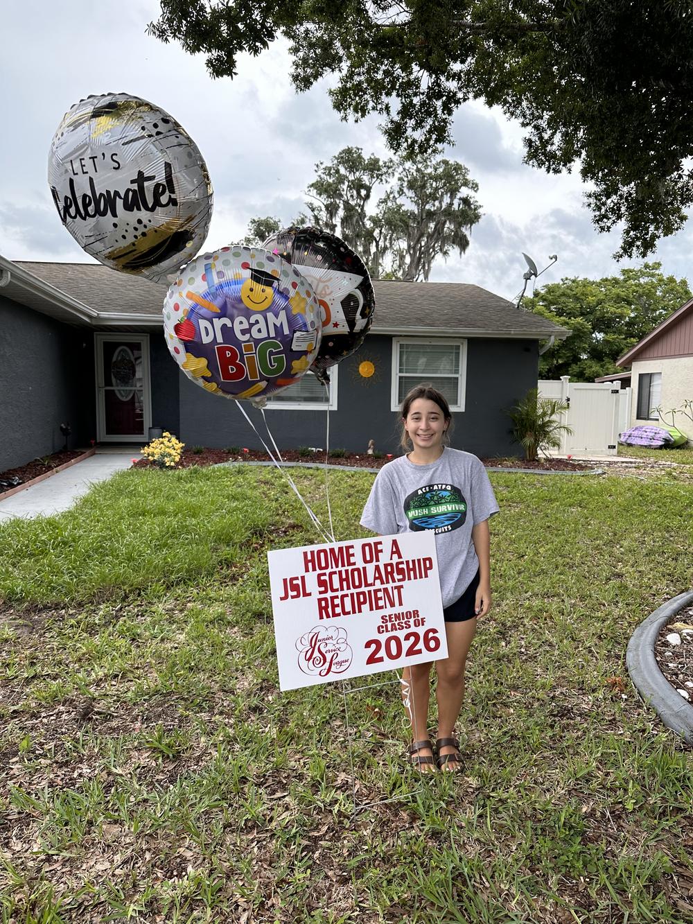 A graduating senior stands beside her Home of a JSL Scholarship Recipient yard sign holding Let's Celebrate and Dream Big balloons