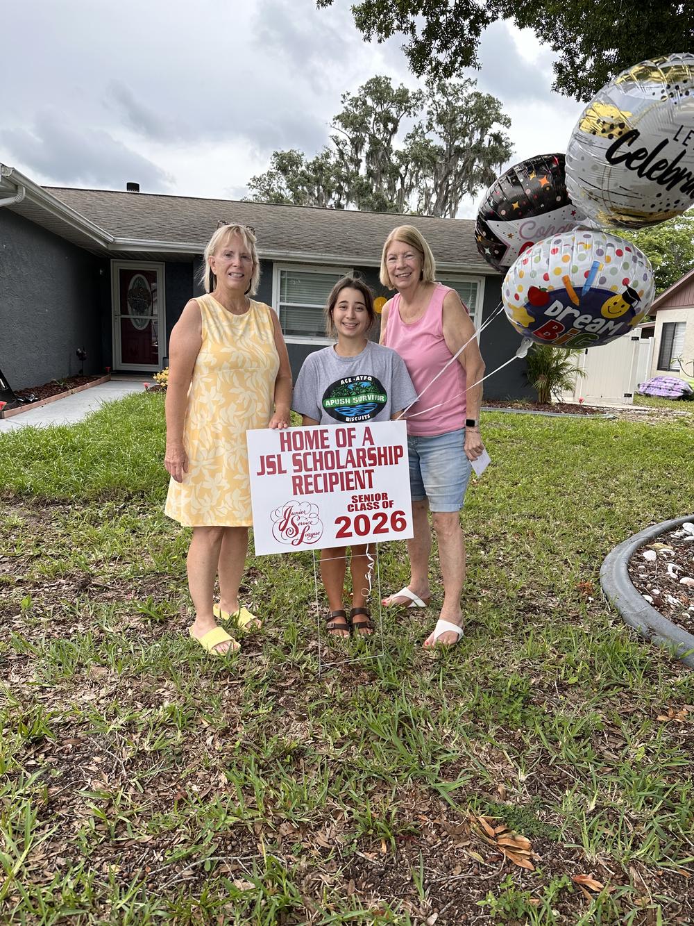 A graduating senior stands with two JSL members behind her Home of a JSL Scholarship Recipient yard sign