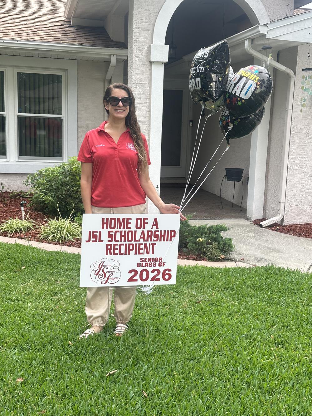 A JSL member in a League polo holds a Home of a JSL Scholarship Recipient yard sign and graduation balloons ready for delivery